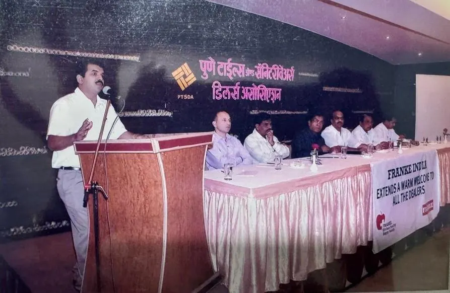 A speaker at the podium during a Pune Tiles and Sanitaryware Dealers Association meeting, with guests seated at the head table